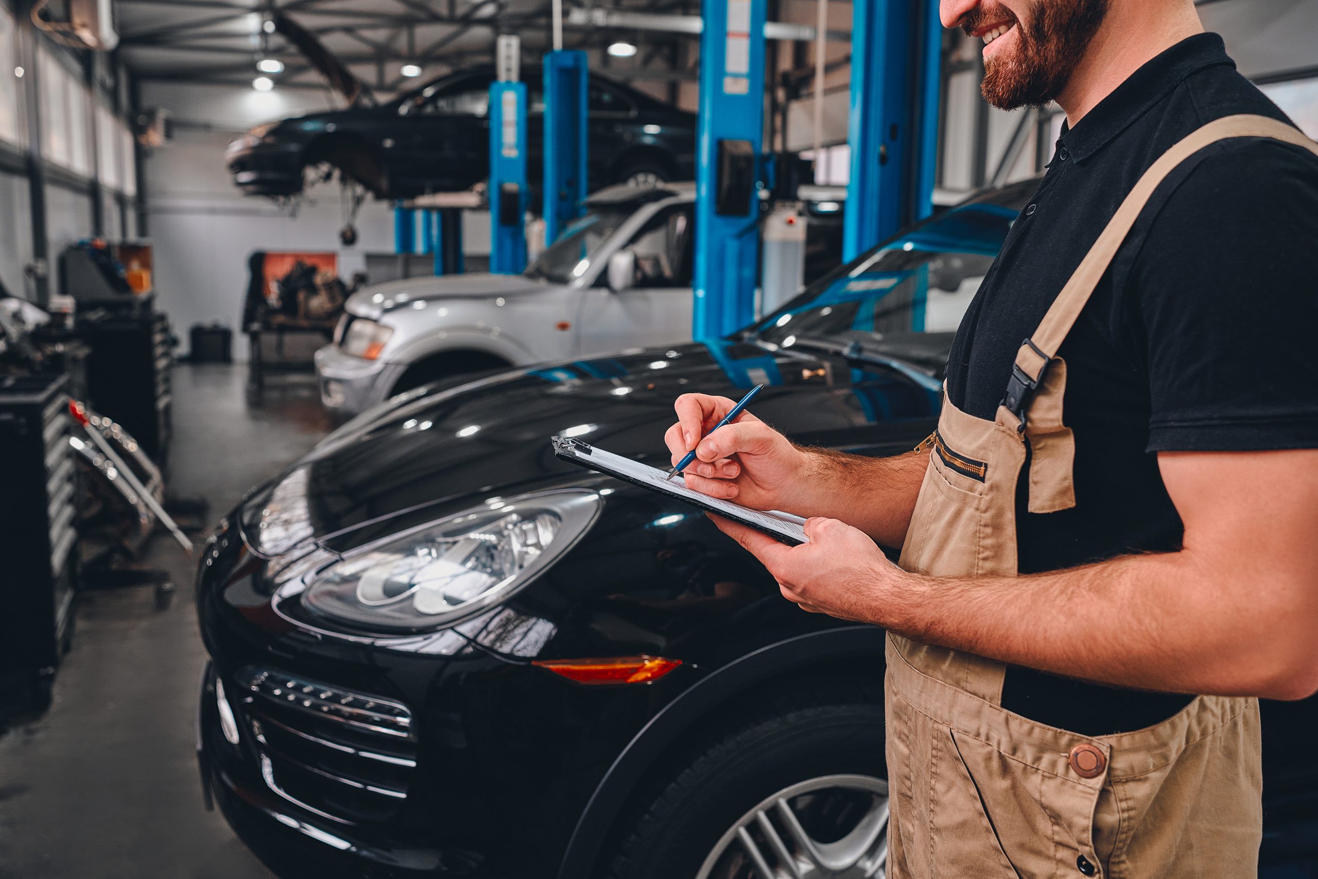 Cropped image of handsome mechanic in uniform making notes while standing in auto service. Side view.