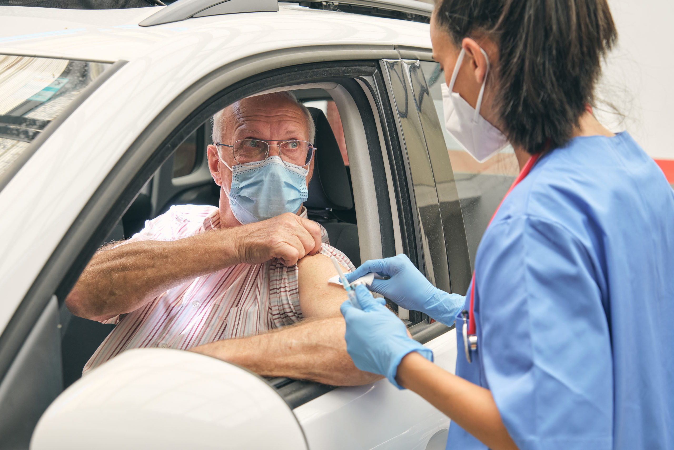 Crop anonymous female physician in uniform touching arm of senior male driver with medical napkin before inoculation in auto