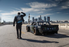 Ken Block facing away and standing next to race car on bridge