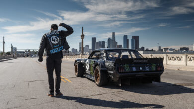 Ken Block facing away and standing next to race car on bridge