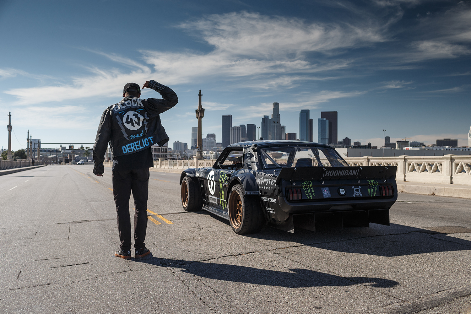 Ken Block facing away and standing next to race car on bridge