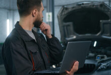 Auto mechanic holding computer and looking concerningly at car engine
