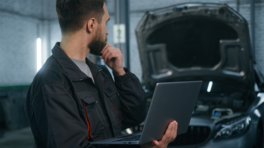 Auto mechanic holding computer and looking concerningly at car engine