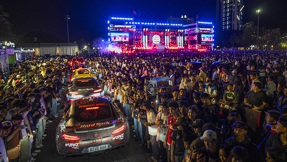 Line of supercars in middle of concert crowd at night with lit stage in background