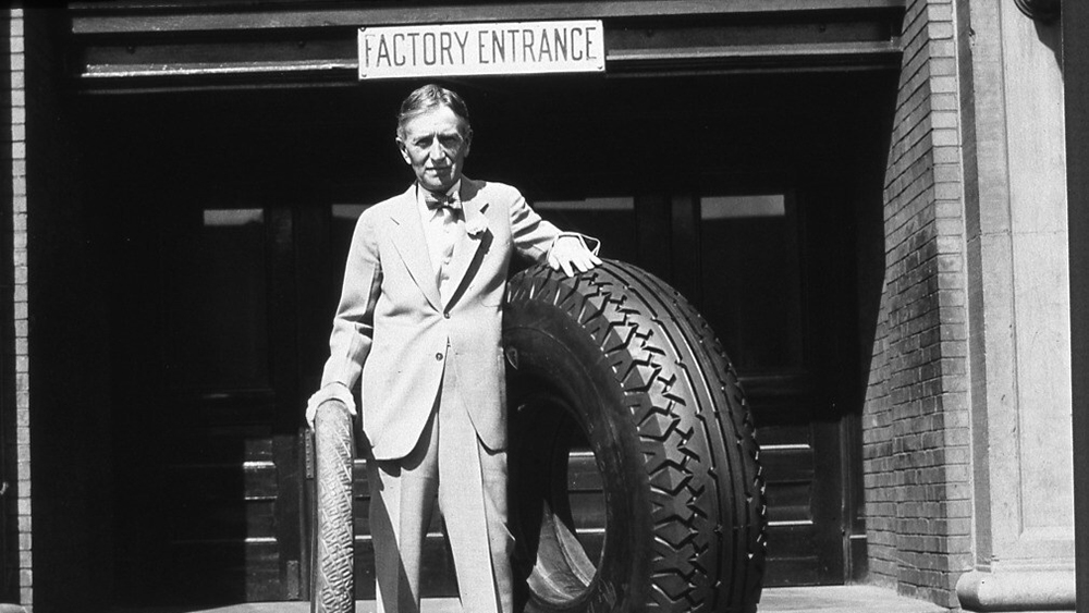 Harvey Firestone Sr. standing at the Akron, Ohio, factory entrance with an original Firestone non-skid tread tire and a large tire, ca. 1926-30.