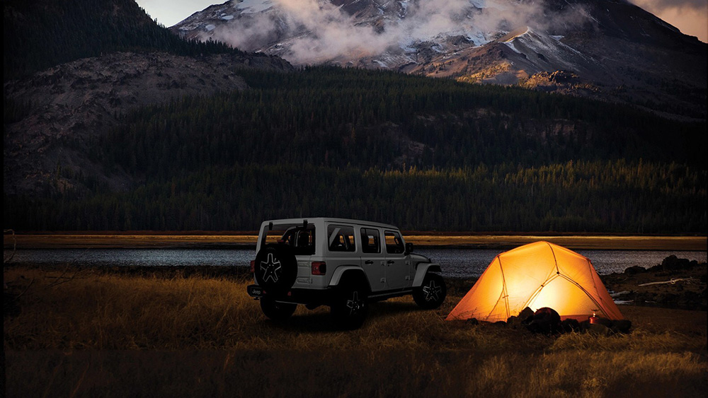 Jeep parked next to lit-up tent with mountains in the background