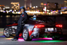 Photo of driver leaning against race car at night with city skyline in background