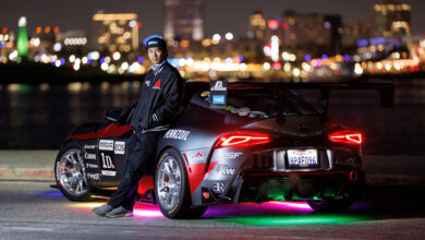 Photo of driver leaning against race car at night with city skyline in background